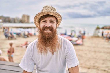 Genç, kızıl saçlı, gülümseyen turist kumsaldaki bankta mutlu bir şekilde oturuyor..