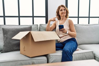 Orta yaşlı bir kadın kanepede oturmuş, teslimat paketini kontrol ediyor ve gülümseyen mutlu ve gururlu birini işaret ediyor. 