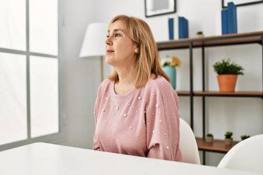 Orta yaşlı beyaz kadın, günlük giysiler içinde evde oturmuş, yan tarafa bakıyor, rahat bir görünüm sergiliyor, kendine güvenen bir gülüşü var.. 