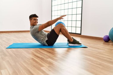 Genç İspanyol adam gülümsüyor. Spor merkezinde top kullanarak karın kasları çalıştırıyor.