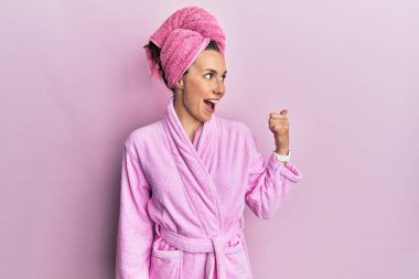 Duş başlığı ve bornoz takan, gülümseyen, gülümseyen ve başparmağıyla yan tarafı gösteren genç sarışın kadın.. 