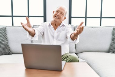 Evde dizüstü bilgisayar kullanan yaşlı bir adam koltukta oturmuş kameraya bakıyor ve sarılmak için kollarını açmış gülümsüyor. Mutluluğu kucaklayan neşeli bir ifade.. 