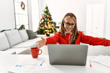 Masada oturan beyaz bir kız Noel ağacının yanında dizüstü bilgisayarını kullanarak sarılıp gülümseyen kameraya bakıyor. Mutluluğu kucaklayan neşeli bir ifade.. 