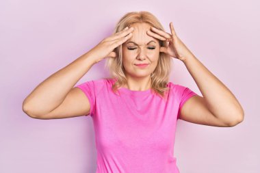 Elleri kafasında, günlük pembe tişört giyen genç sarışın kadın, stres yüzünden baş ağrısı çekiyor. migren ağrısı. 