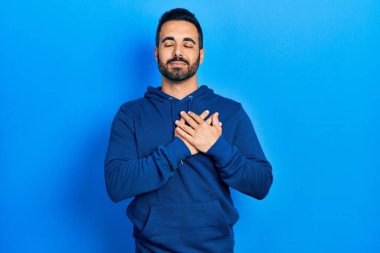 Sakallı, sıradan tişörtlü, göğsünde elleri göğsünde, yüzünde minnet dolu bir jest ile gülümseyen yakışıklı bir İspanyol. sağlık kavramı. 