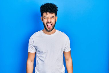 Sakallı, sıradan beyaz tişörtlü, dilini dışarı çıkaran komik ifadeli genç bir Arap. duygu kavramı. 