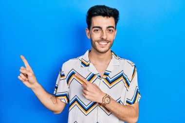 Gündelik kıyafetler giyen genç İspanyol adam gülümseyip kameraya bakıyor ve iki eliyle ve parmaklarıyla kenarı işaret ediyor.. 