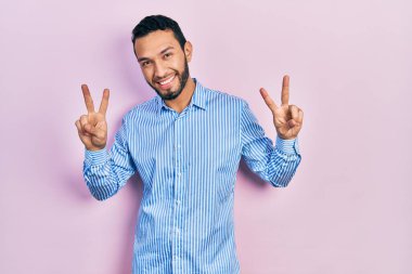 Sakallı, sıradan mavi gömlekli, gülümseyen, parmaklarını zafer işareti yaparken gösteren İspanyol bir adam. İki numara.. 