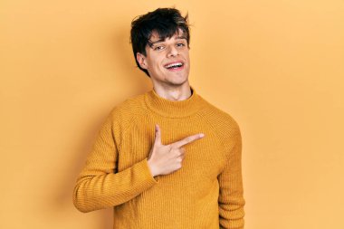 Yakışıklı hippi genç adam gündelik sarı süveter giyiyor. Yüzünde gülücük var. El ve parmaklarıyla mutlu ve doğal bir ifadeyle yüzünü gösteriyor. 