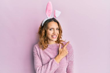 Paskalya tavşanı kulakları takan, neşeli gülümseyen, eli ve parmağı yanda genç, beyaz bir kadın. 
