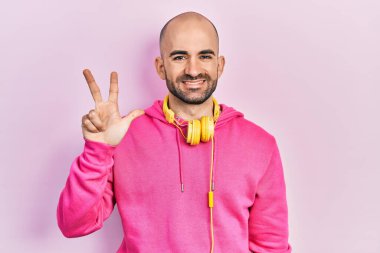 Spor kıyafetleri giyen ve kulaklık takan genç kel bir adam üç numarayı gösterip işaret ederken kendinden emin ve mutlu bir şekilde gülümsüyor.. 