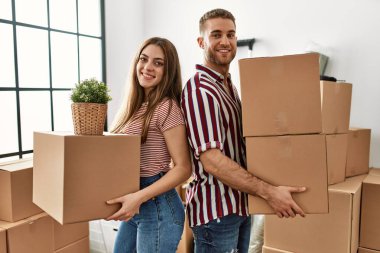 Genç beyaz çift mutlu bir şekilde gülümsüyor. Yeni evlerinde karton kutu tutuyorlar..