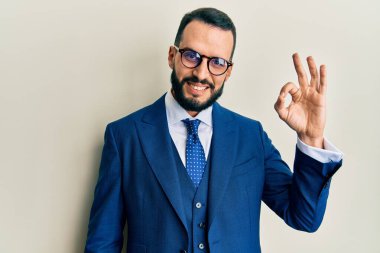 Sakallı, takım elbiseli ve kravatlı genç adam gülümsüyor. El ve parmaklarıyla tamam işareti yapıyor. başarılı ifade. 