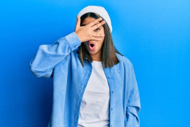 Şirin yün şapkalı genç Latin bir kadın. Şoka girip yüzünü ve gözlerini kapatıyor. Utanmış bir ifadeyle parmaklarının arasından bakıyor.. 