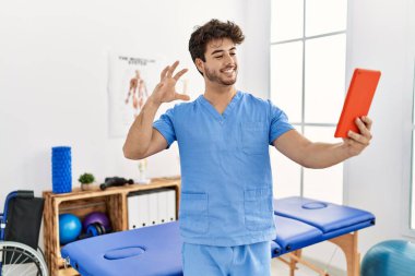 Young hispanic man wearing physio therapist uniform having video call at clinic