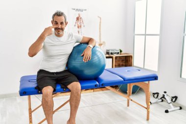 Orta yaşlı İspanyol bir adam ağrıyı dindirme kliniğinde elinde pilates topuyla el ve burnu işaret ediyor, neşeli bir şekilde gülümsüyor. güzellik konsepti 