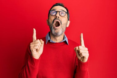 Orta yaşlı İspanyol bir adam gündelik giysiler ve gözlük takıyor şaşkın şaşkın yukarı bakıyor ve parmaklarıyla işaret ediyor ve kollarını kaldırıyor.. 