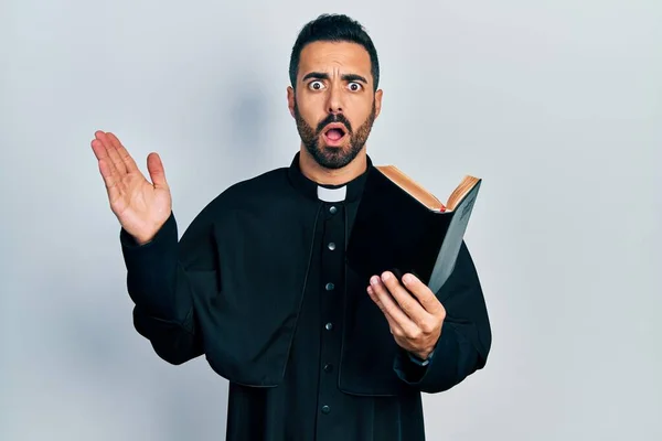 Pohledný Hispánský Kněz Vousy Držící Bibli Křesťanský Kříž Vyděšený Šokovaný — Stock fotografie