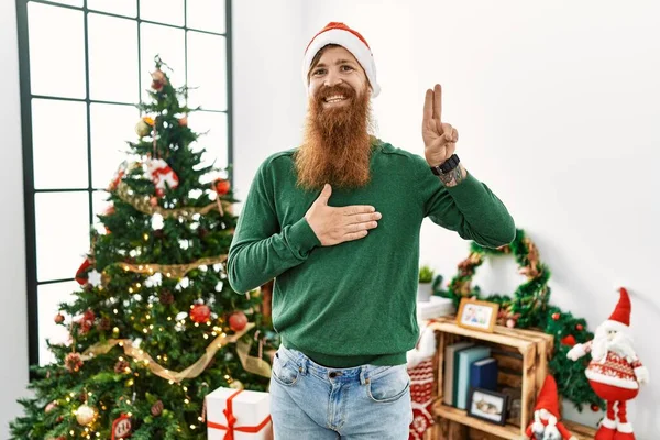 红头发男人留着长胡子 头戴圣诞帽 头戴圣诞礼帽 头戴圣诞礼帽 面带微笑 双手托着胸膛 手指头高举 宣誓效忠 — 图库照片