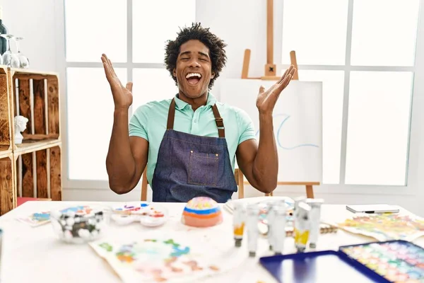 若いアフリカ系アメリカ人の男性がアートスタジオのテーブルの上に座って 腕を上げて興奮して叫んで目を閉じて成功のために狂気と狂気を祝う 勝者のコンセプト — ストック写真