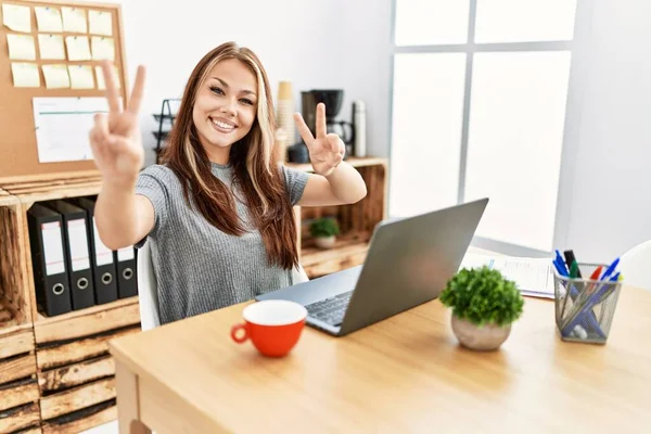 年轻的黑发女人在办公室里工作 带着笔记本电脑微笑看着摄像机 手指在做胜利的标志 第二点 — 图库照片