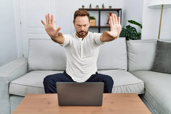 Middle age man using laptop at home doing stop gesture with hands palms, angry and frustration expression