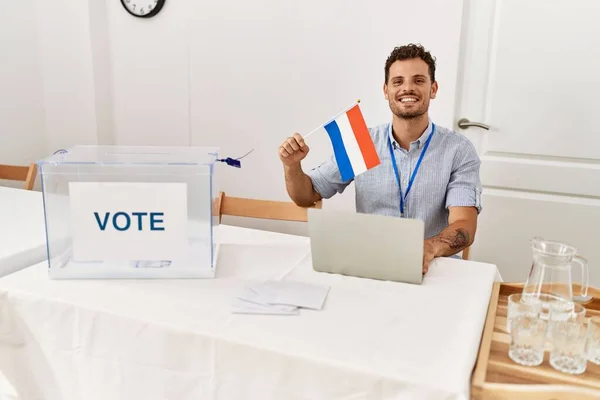 Joven Hispano Sonriendo Confiado Sosteniendo Bandera Holandesa Trabajando Colegio Electoral —  Fotos de Stock