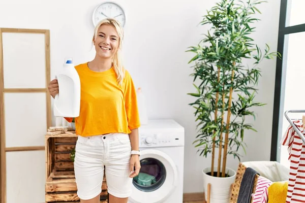 Mulher Branca Jovem Fazendo Lavandaria Segurando Garrafa Detergente Olhando Positivo — Fotografia de Stock