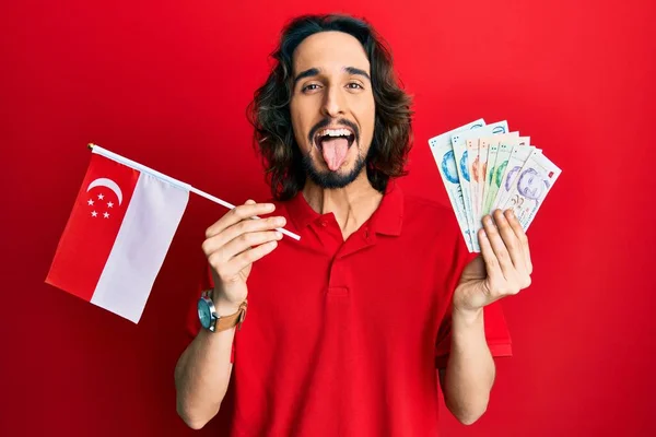 Joven Hombre Hispano Sosteniendo Bandera Singapur Dólares Sacando Lengua Feliz —  Fotos de Stock