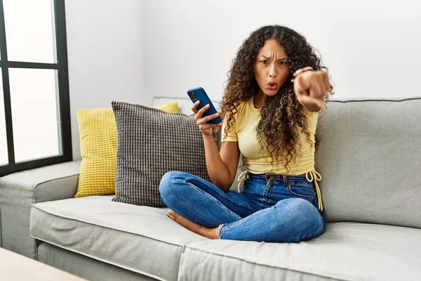 カメラに不満と不満を指しているスマートフォンを使用して自宅でソファに座っている美しいヒスパニック系の女性 怒っているとあなたと激怒 — ストック写真