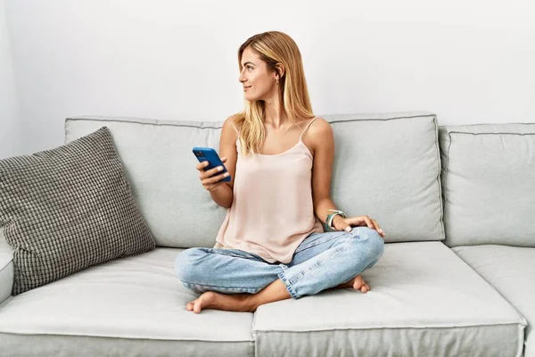 金发美女坐在家里沙发上 用智能手机向旁边看去 用自然的脸和自信的微笑放松自己的形象 — 图库照片