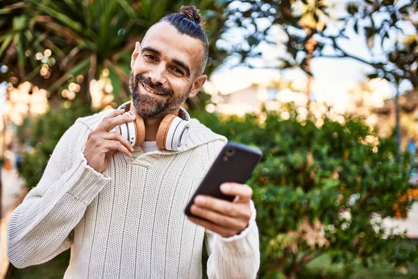 髭を生やしたハンサムな中年男性は スマートフォンやヘッドフォンを使用して幸せと自信を持って屋外に立っています — ストック写真