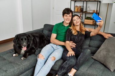 Kafkasyalı genç bir çift akıllı telefonun başında evde köpekle kanepede oturmuş selfie çekiyor..
