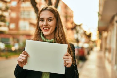 Genç sarışın kız mutlu bir şekilde gülümsüyor şehirde boş bir pankart tutuyor..