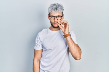 Modern boyalı saçlı, beyaz tişörtlü, gözlüklü ağzı ve dudakları parmakları fermuarlı İspanyol genç bir adam. gizli ve sessiz, tabu konuşması 