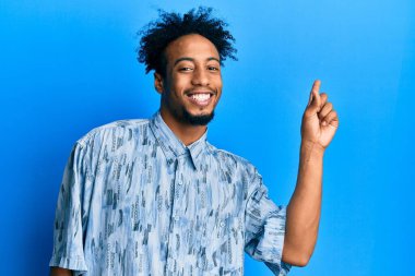 Sakallı, sıradan giyinmiş, yüzünde kocaman bir gülümseme olan, eli ve parmağı kameraya bakan Afro-Amerikalı genç bir adam.. 