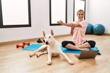 Genç beyaz kadın spor merkezinde köpekle internetten esneme dersi alıyor.