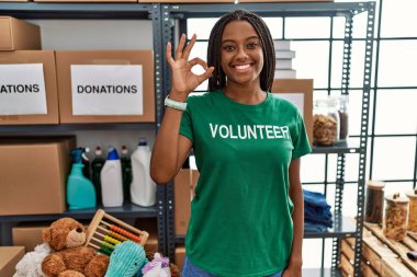 Bağışlarda gönüllü tişört giyen genç Afro-Amerikalı kadın gülümsüyor ve el ve parmaklarıyla tamam işareti yapıyor. başarılı ifade. 