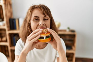 Beyaz kadın klasik hamburger yiyor. Evdeki masada oturuyor.