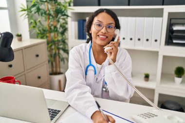 Doktor üniforması giyen genç Latin kadın telefonla konuşuyor. Klinikteki panoya yazı yazıyor.