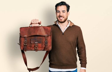 Deri çanta giyen genç İspanyol adam pozitif ve mutlu görünüyor. Dişlerini gösteren kendine güvenen bir gülümsemeyle ayakta duruyor ve gülümsüyor. 