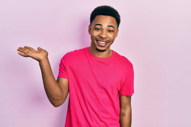 Casua tişörtü giyen genç Afro-Amerikan adam mutlu bir şekilde gülümsüyor ve avuç içiyle kameraya bakıyor.. 