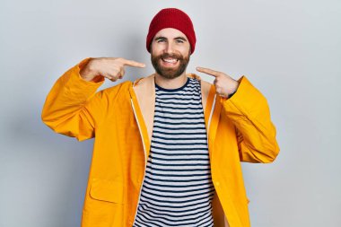 Sakallı, sarı yağmurluk giyen neşeli gülümseyen ve parmaklarıyla ve ağzıyla işaret eden beyaz bir adam. diş sağlığı kavramı. 