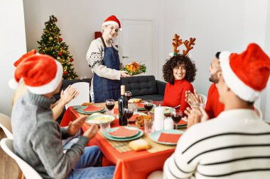 Bir grup insan el çırpıyor ve masada oturuyor. Ayakta duran ve elinde kavrulmuş hindi tutan kadın Noel 'i evde kutluyor..