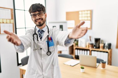 Doktor üniforması giyen ve steteskop takmış İspanyol bir adam kameraya bakıp sarılmak için gülümsüyor. Mutluluğu kucaklayan neşeli bir ifade.. 