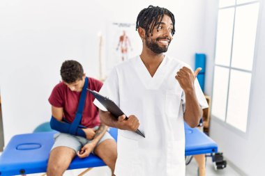 Kırık kollu, gülümseyen ve baş parmağıyla yüzünü gösteren bir adamla birlikte ağrı kesici kliniğinde çalışan İspanyol genç bir adam.. 