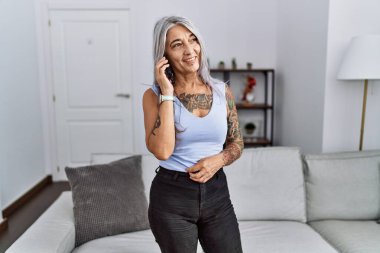 Orta yaşlı, kır saçlı, gülümseyen, kendine güvenen bir kadın evde akıllı telefondan konuşuyor.