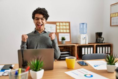 Young hispanic man wearing business style sitting on desk at office excited for success with arms raised and eyes closed celebrating victory smiling. winner concept. 