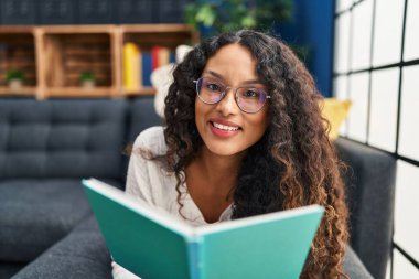 Evdeki kanepede kitap okuyan genç Latin kadın.