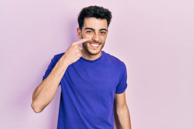 Gündelik tişört giyen genç İspanyol adam, el parmağı ile yüzünü ve burnunu gösteriyor, neşeli bir şekilde gülümsüyor. güzellik konsepti 